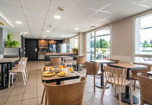 A modern dining area with tables and chairs, a kitchen in the background, and large windows offering a view of the outdoors.