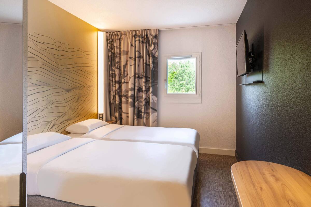 Una habitación de hotel con dos camas, una ventana, un banco de madera y un televisor de pared.