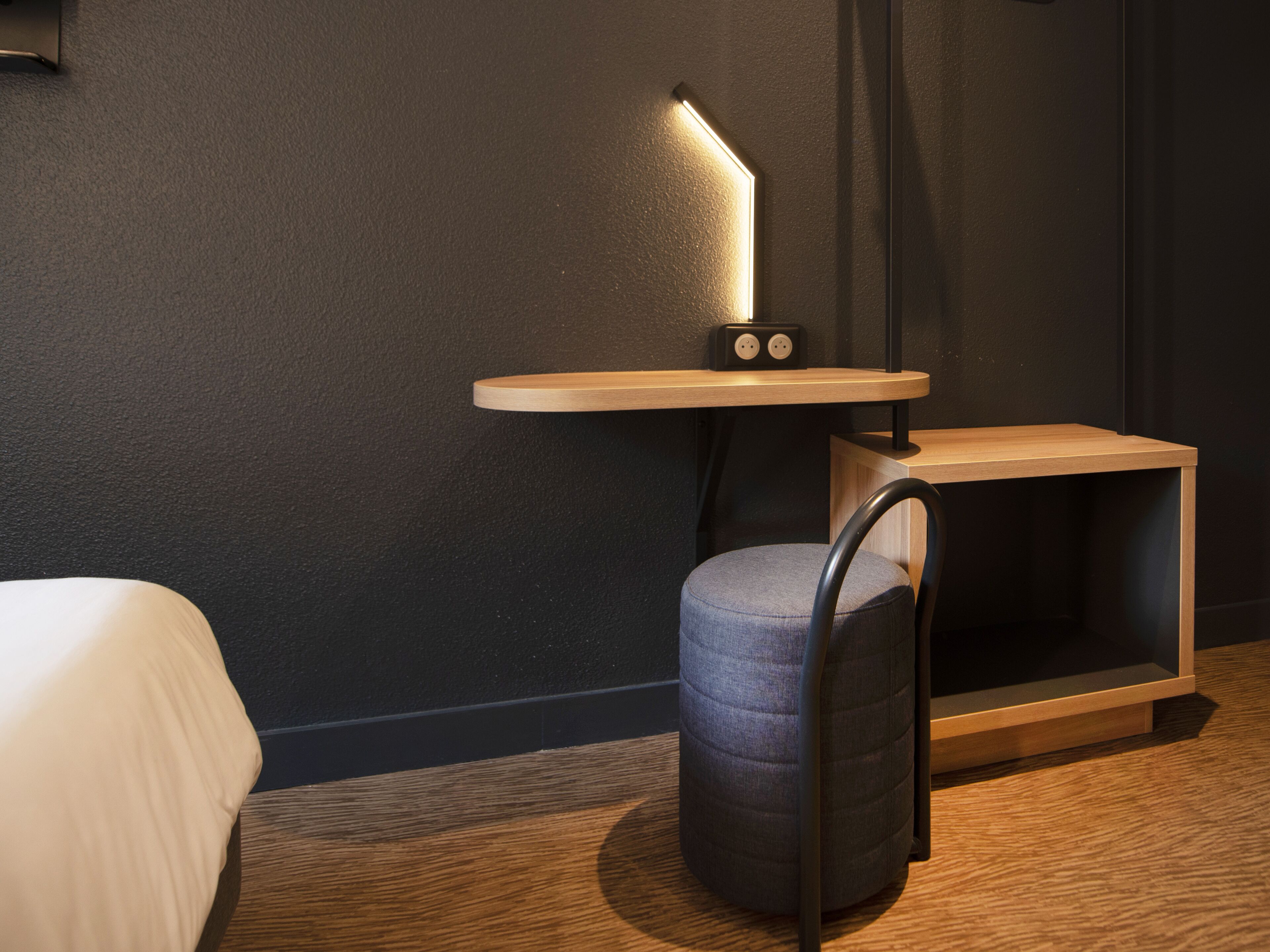 A modern hotel room with a wooden desk, a chair, a bedside table, and a lamp.