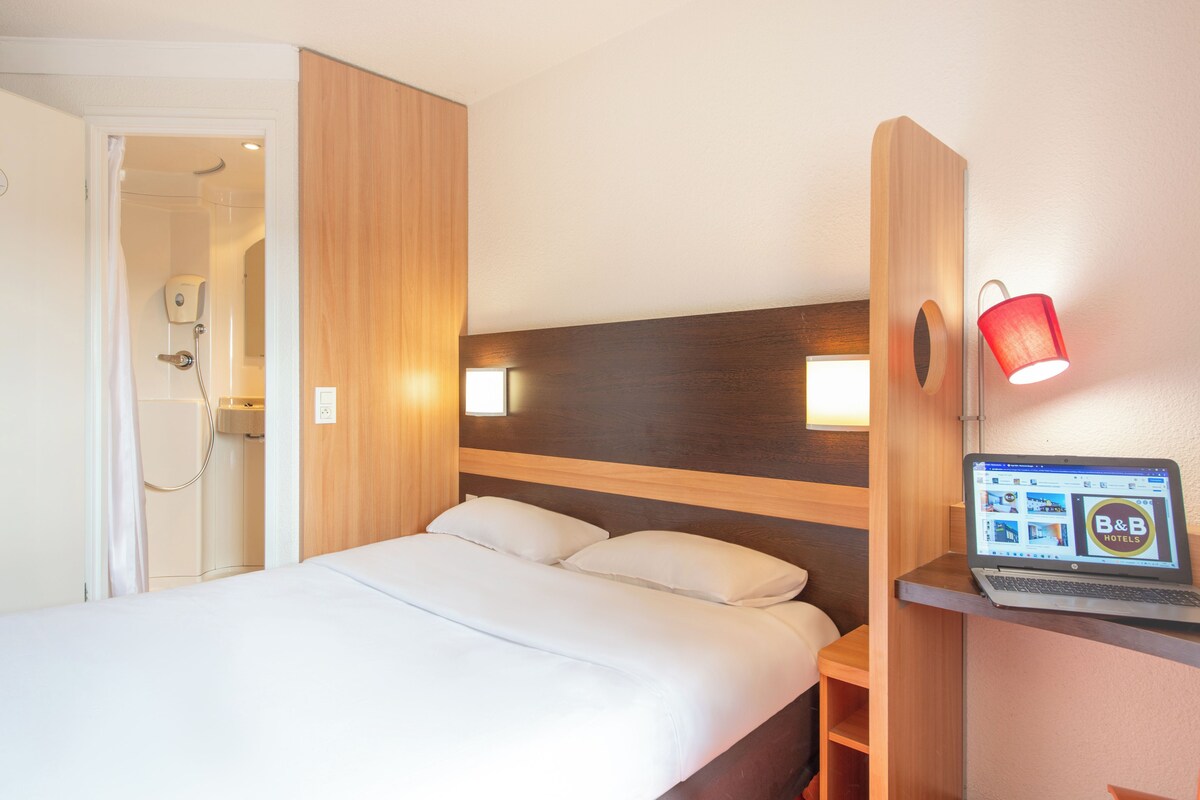 A hotel room with a bed, a wooden headboard, a laptop on a desk, and a red lamp.