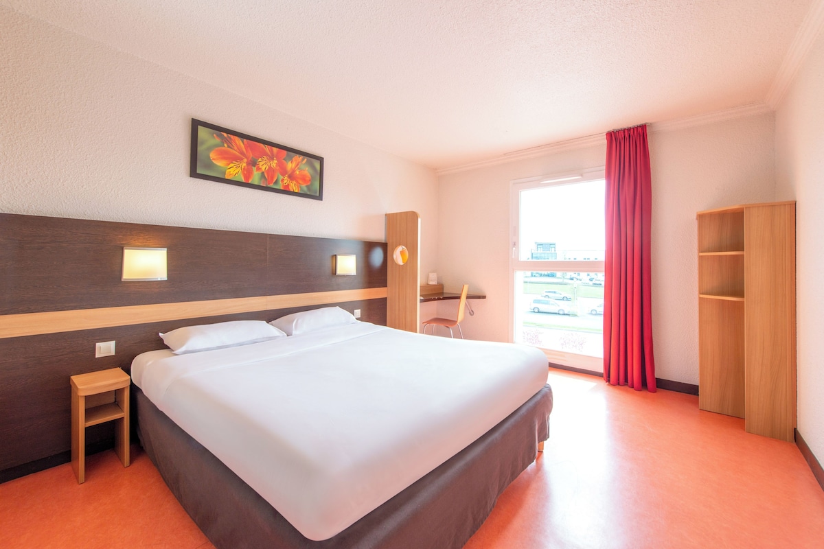 A hotel room with a bed, bedside table, lamp, chair, and a view of the outside.