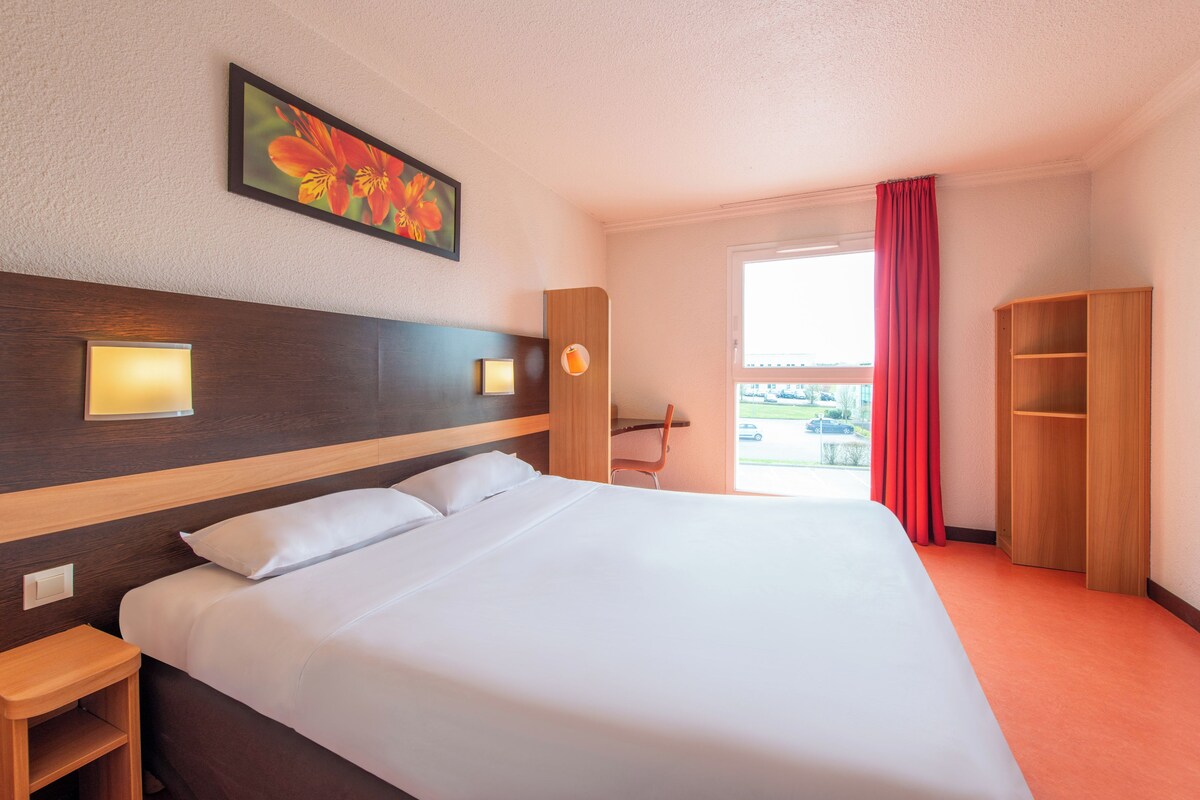 Habitación de hotel con cama, mesitas de noche, escritorio, silla, armario, ventana con cortinas y un cuadro en la pared.