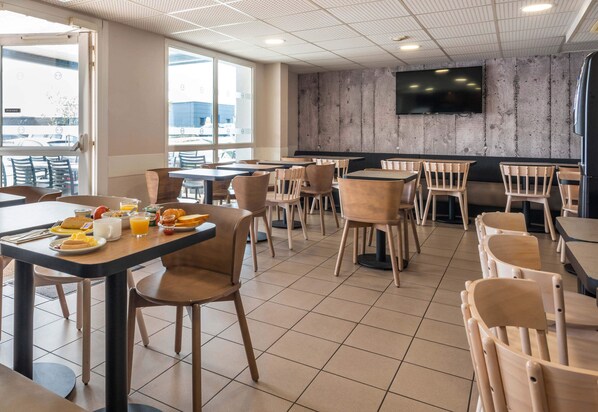 A modern dining area with tables and chairs, a flat-screen TV on the wall, and large windows providing a view of the outside. - B&B HOTEL Cholet Nord (Cholet)