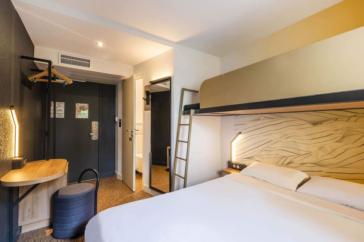 Habitación de hotel con literas, una mesita de noche, una lámpara, un perchero y un taburete pequeño.