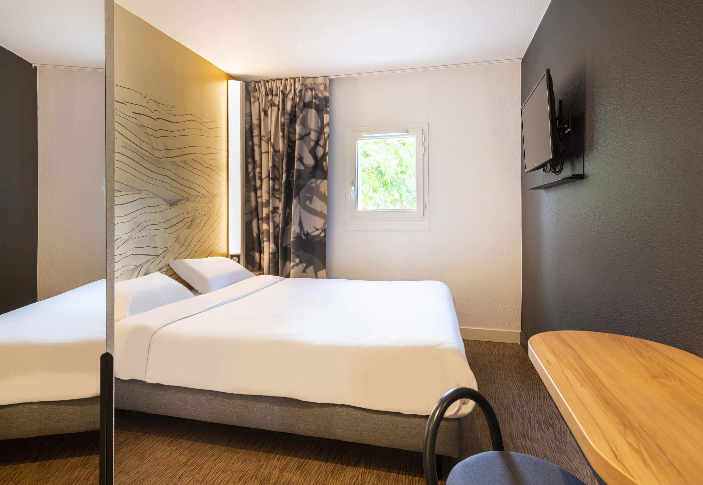 A hotel room with two beds, a small window, a TV mounted on the wall, and a wooden table.