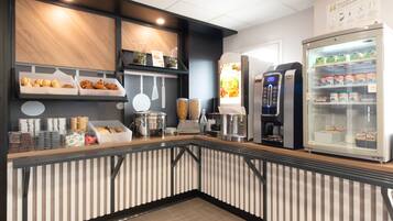 Un espace de petit-déjeuner en libre-service comprenant une machine à café, un présentoir réfrigéré et divers produits alimentaires.