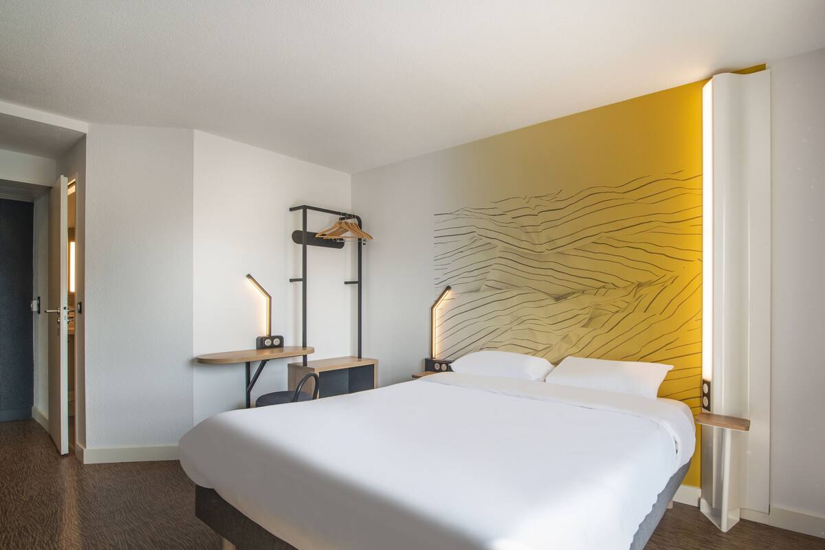 Una habitación de hotel moderna con una cama grande, mesitas de noche, una lámpara fijada a la pared y una puerta que da a otra habitación.