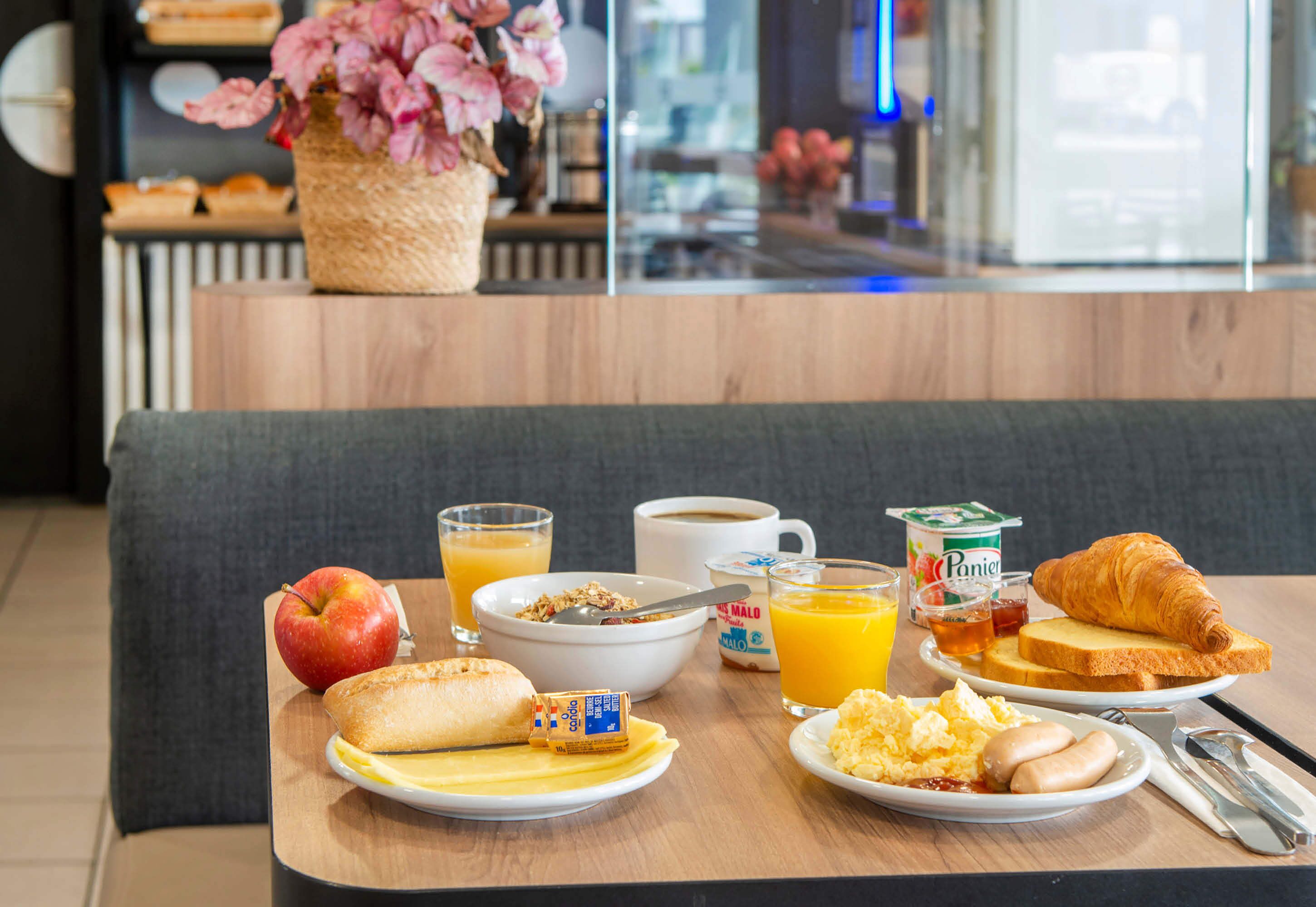 A breakfast table with various food items and drinks, a bowl of cereal, a croissant, an apple, and a glass of orange juice.