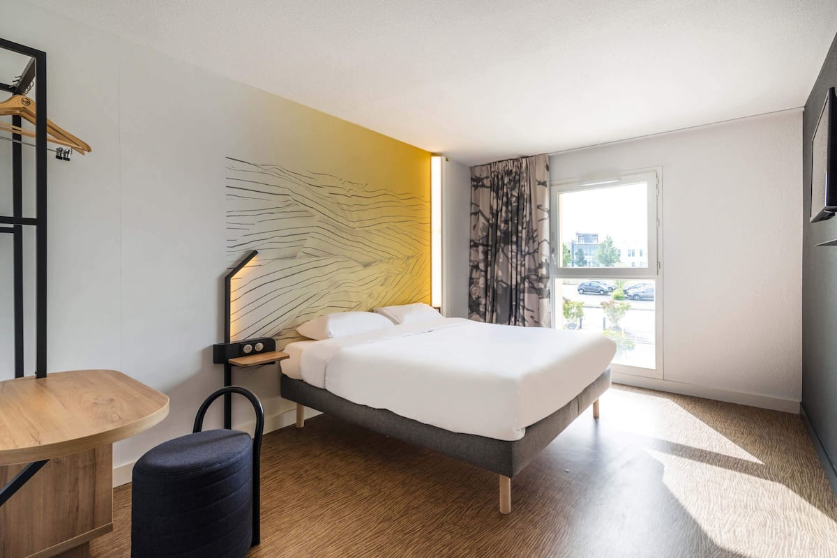 Una habitación de hotel moderna con una cama grande, un escritorio de madera, una silla, una mesita de noche y vistas a la ciudad a través de una ventana.