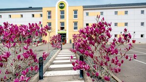 Een B&B hotel met een geel gevel, een centrale ingang, en bloeiende roze bomen langs het pad.