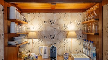 Un área de cocina con estantes de madera que sostienen platos, tazas y vasos, una tetera, una cafetera y un dispensador de agua.