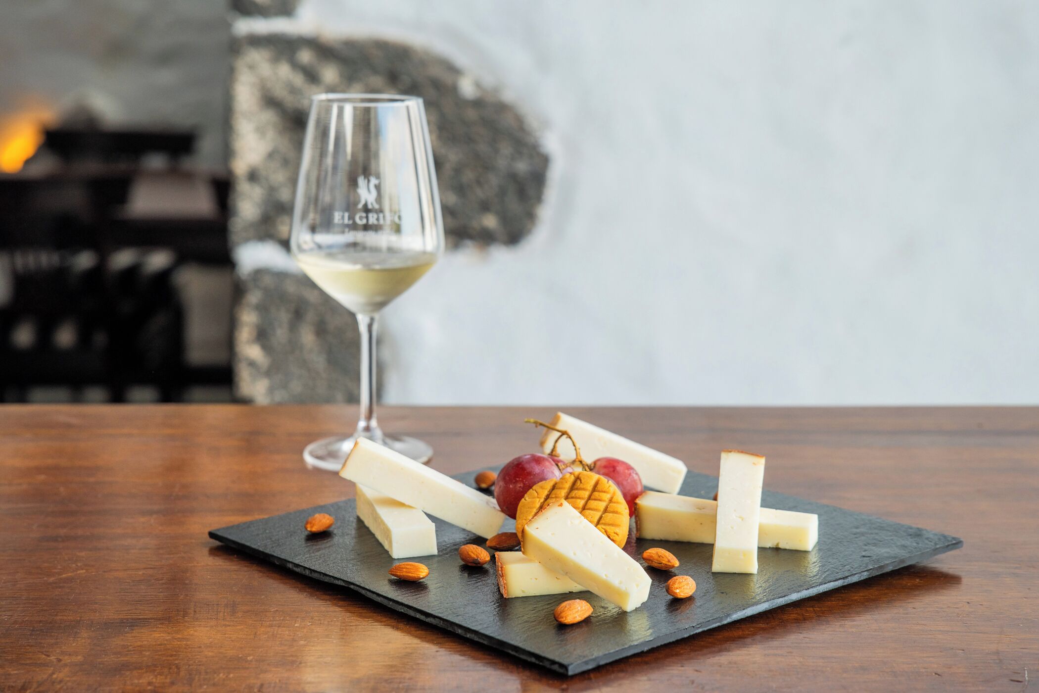 A cheese board with various cheeses, almonds, and grapes on a slate board, accompanied by a wine glass.