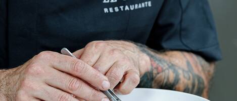 A person wearing a black t-shirt with "EL GRIFO RESTAURANTE" text, sitting at a table with a white bowl containing food.