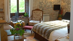 A bedroom with a stone wall, wooden beams, a bed, a chair, a table, a window, and a TV. - Hotel-SPA El Molino de Alcuneza (Siguenza)