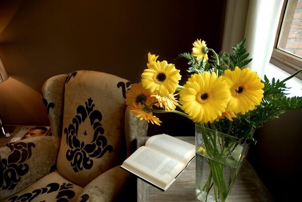 A chair with a patterned cushion, a clear vase with yellow flowers, and an open book on a table.