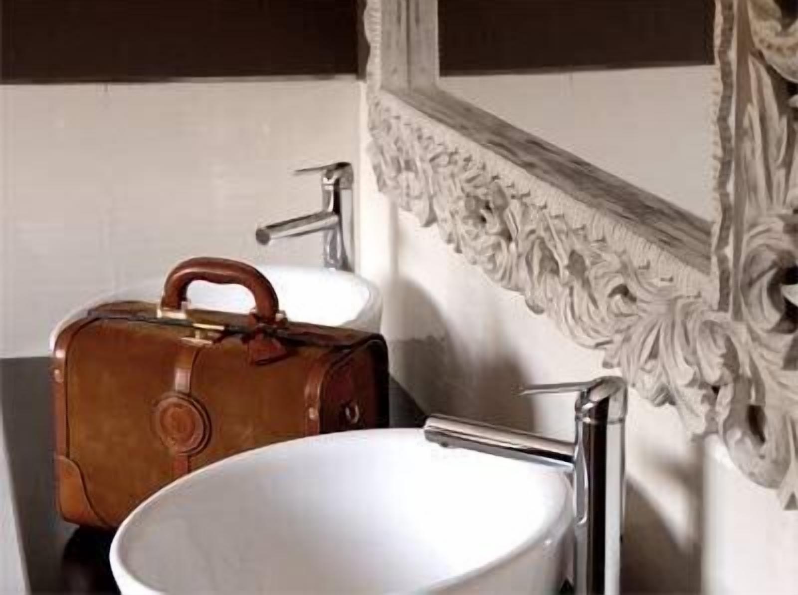 Un baño con un lavabo blanco, un bolso de cuero marrón y un espejo con detalles ornamentales.