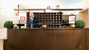 Un comptoir d’accueil avec un homme assis derrière, un écran numérique et une étagère contenant des livres et un panneau.