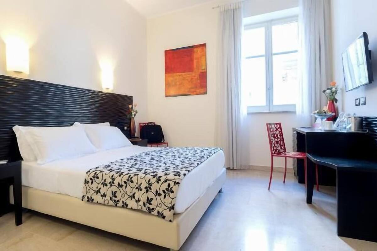 Une chambre d’hôtel avec un lit, une table de chevet, une lampe, une chaise, un bureau et un tableau au mur.