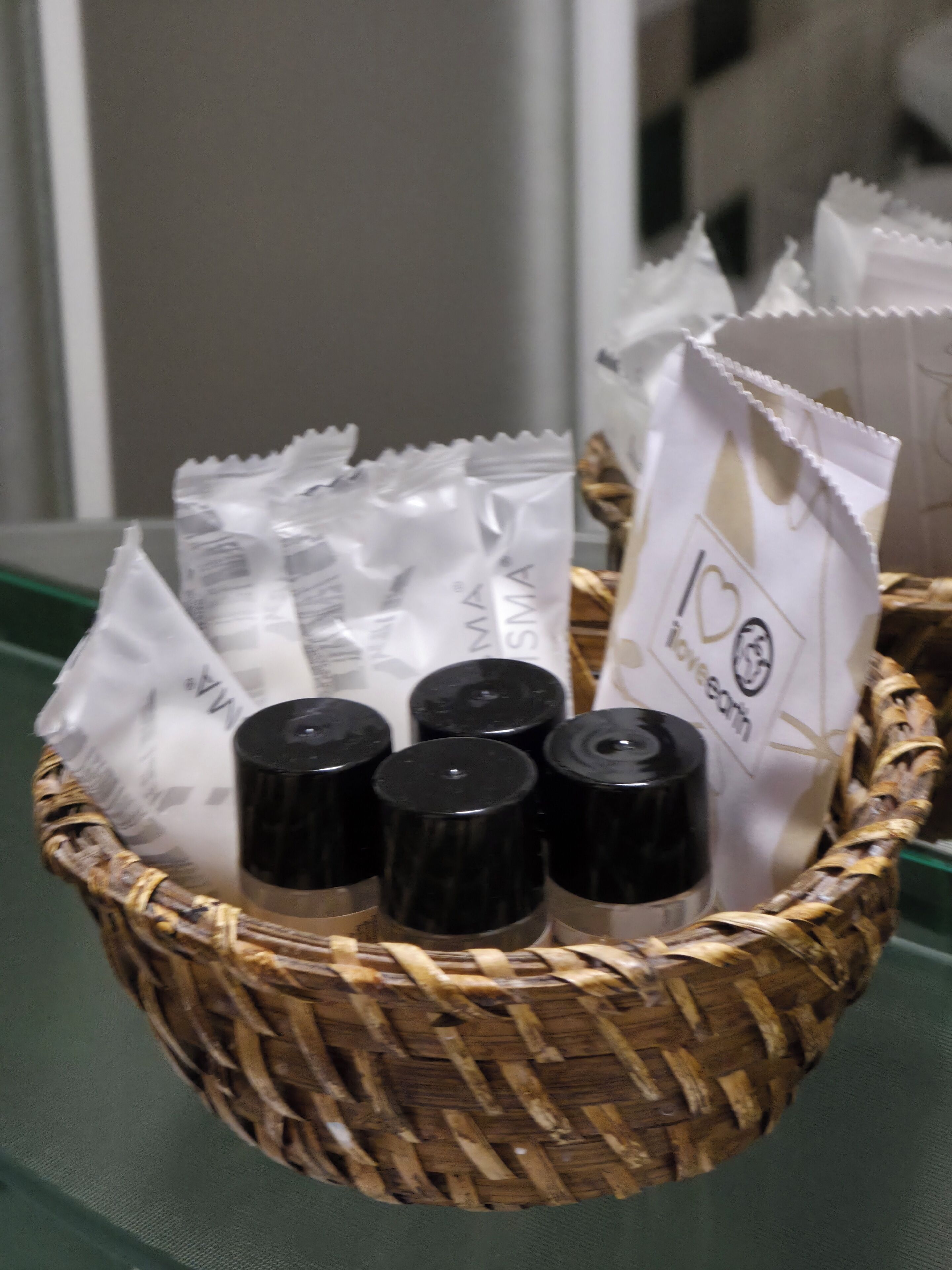 Un panier en osier contenant des récipients noirs et des paquets de papier blancs, le tout posé sur une surface verte.