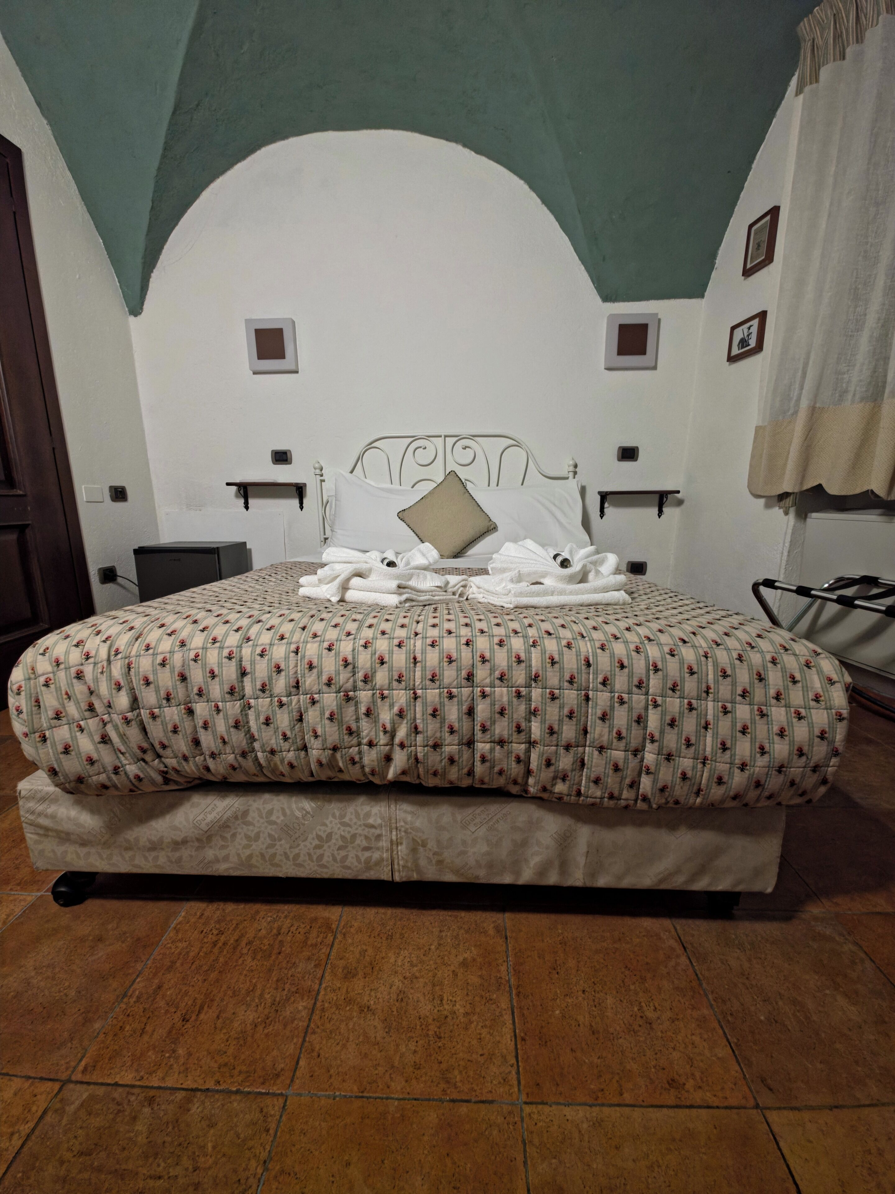 A bed with a floral-patterned bedspread, a headboard, and a wall-mounted shelf with a small picture frame.