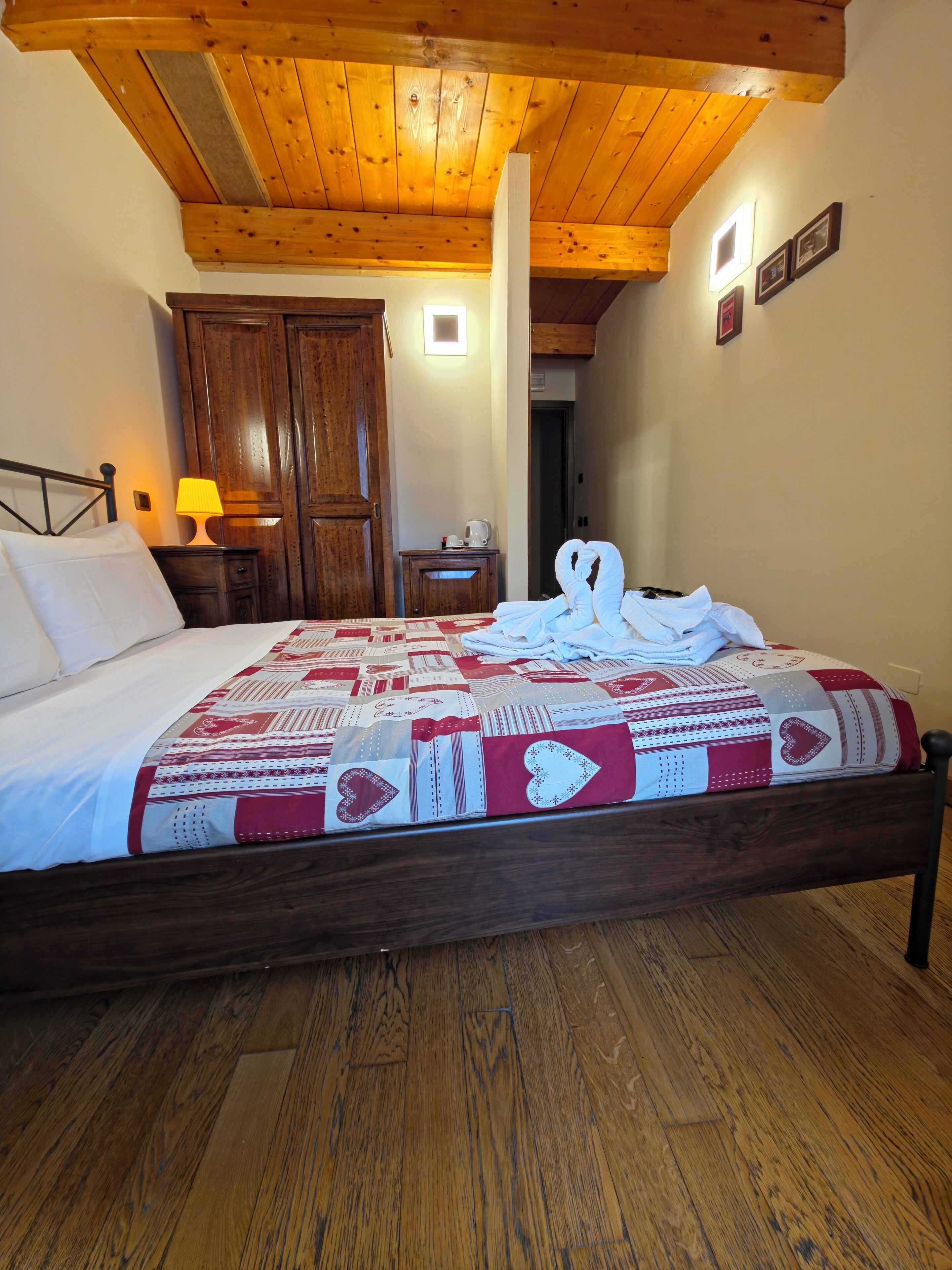 Une chambre avec un plafond en bois, un lit recouvert d’une couvre-lit à motifs, une table de chevet avec une lampe et une armoire en bois.