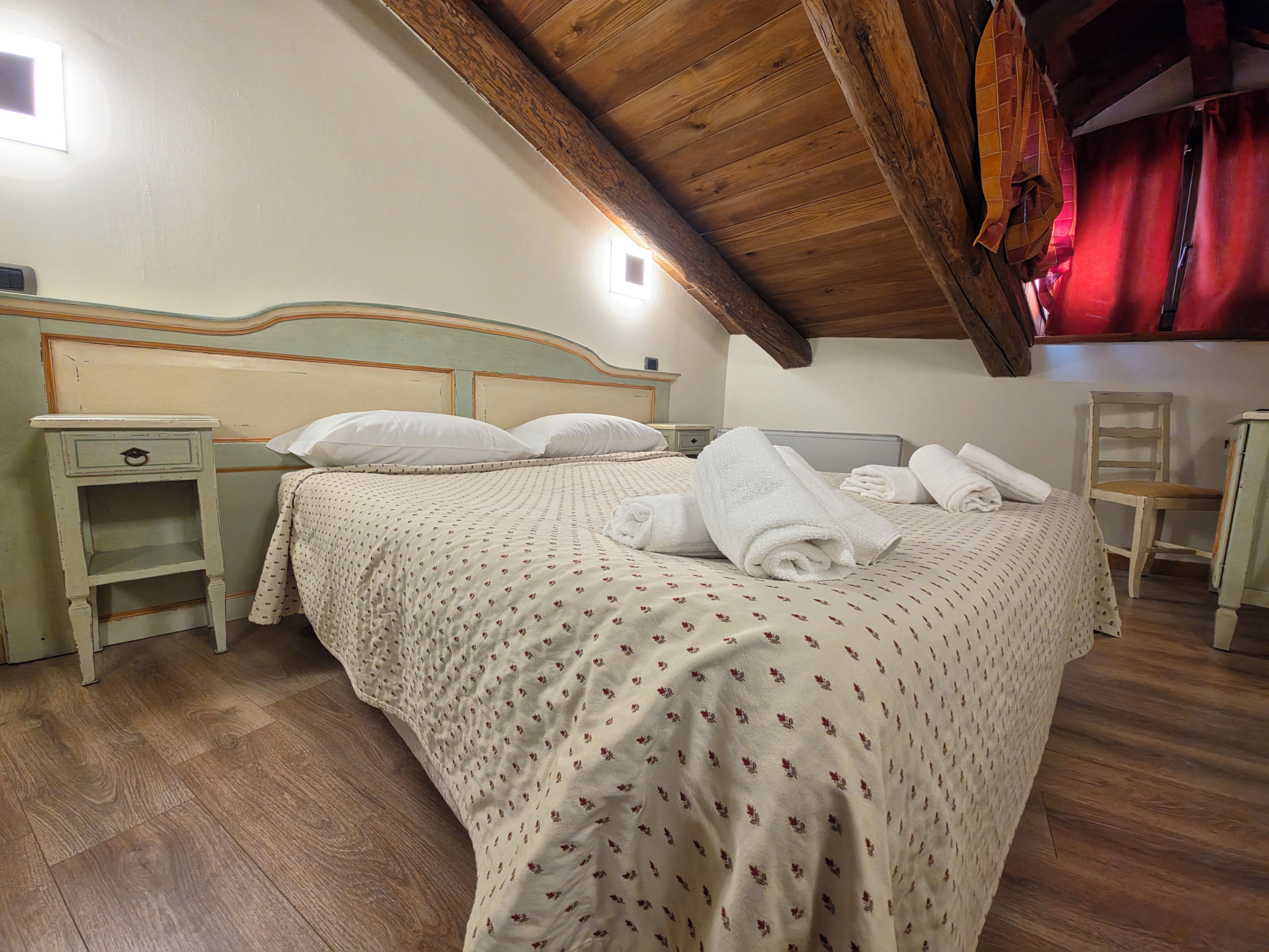 Un dormitorio con techo de madera, una cama con colcha floral, una mesita de noche, una silla y una cortina roja.