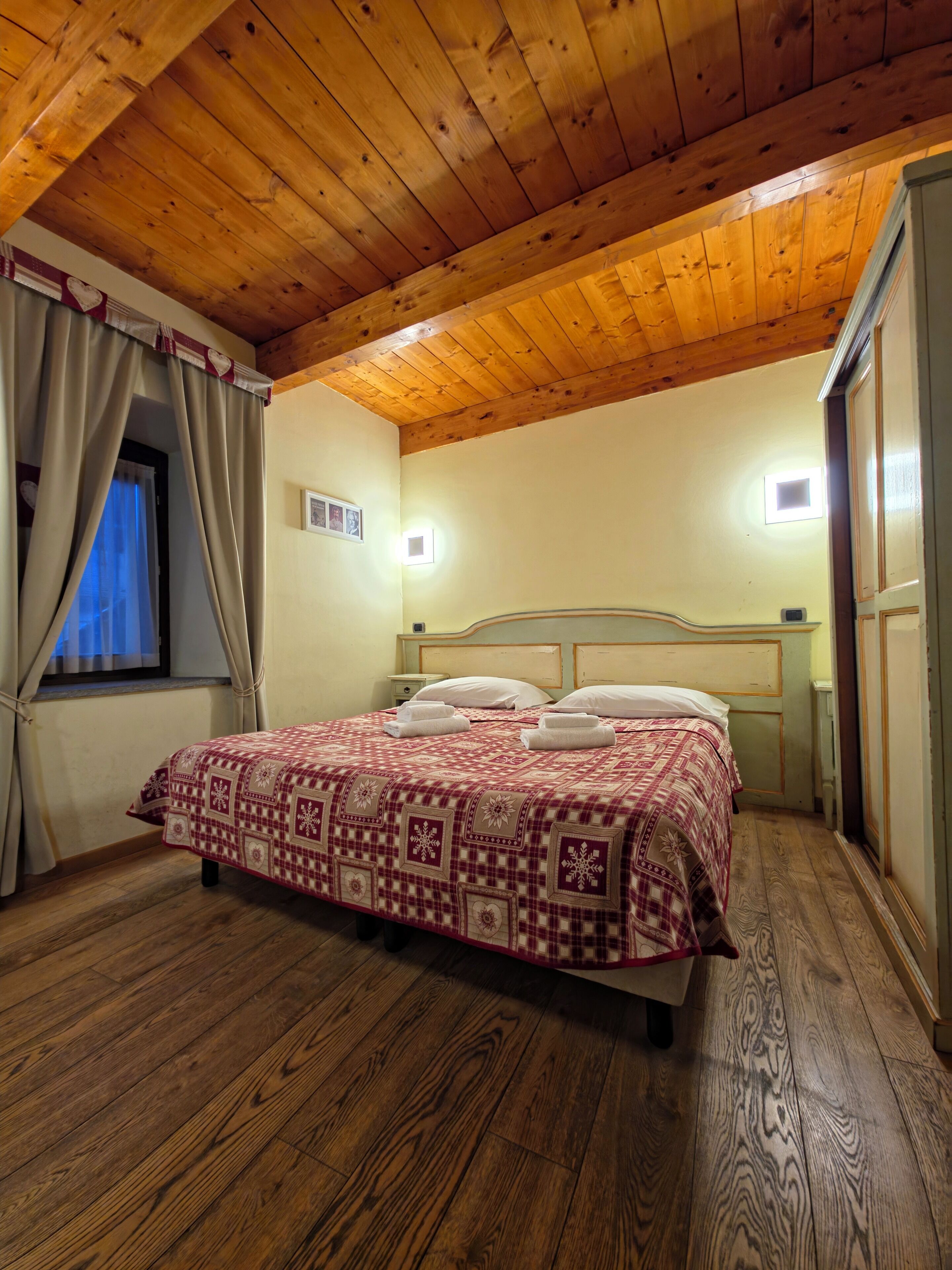 Une chambre avec un plafond en bois, un lit recouvert d’une couvre-lit à motifs, une table de chevet et une fenêtre avec des rideaux.
