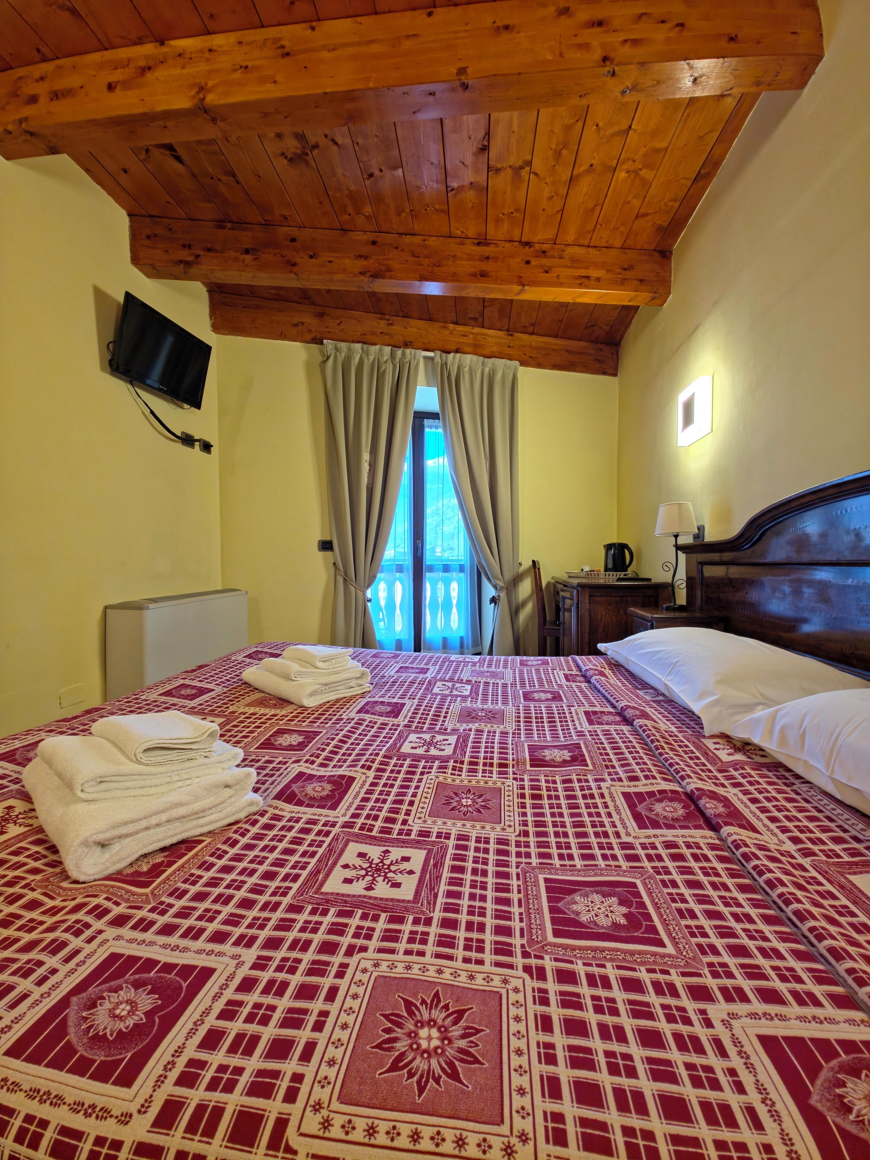 Habitación de hotel con techo de madera, una cama con colcha estampada, un televisor montado en la pared y un balcón visible a través de la ventana.