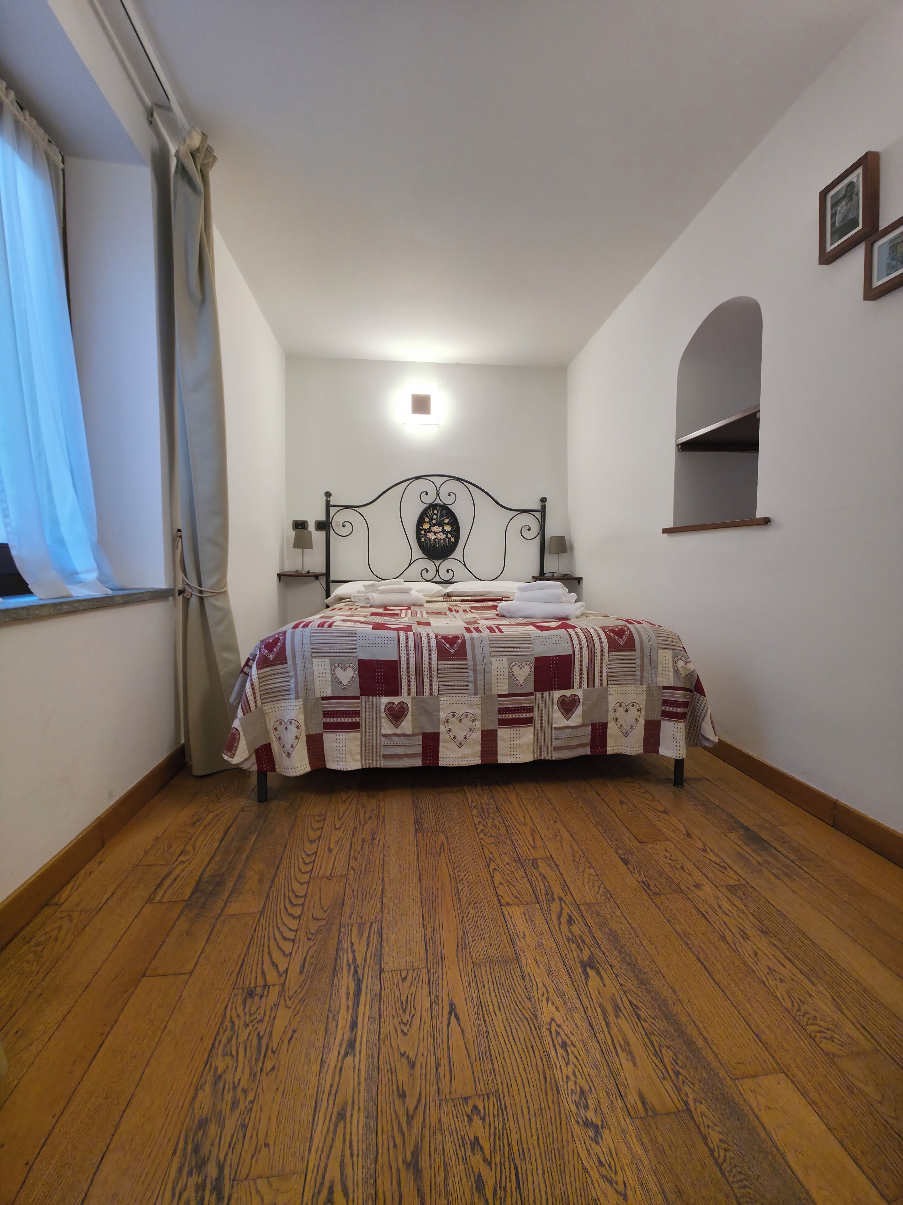 A bedroom with a bed, wooden flooring, a window with curtains, a bedside lamp, and a wall-mounted shelf.