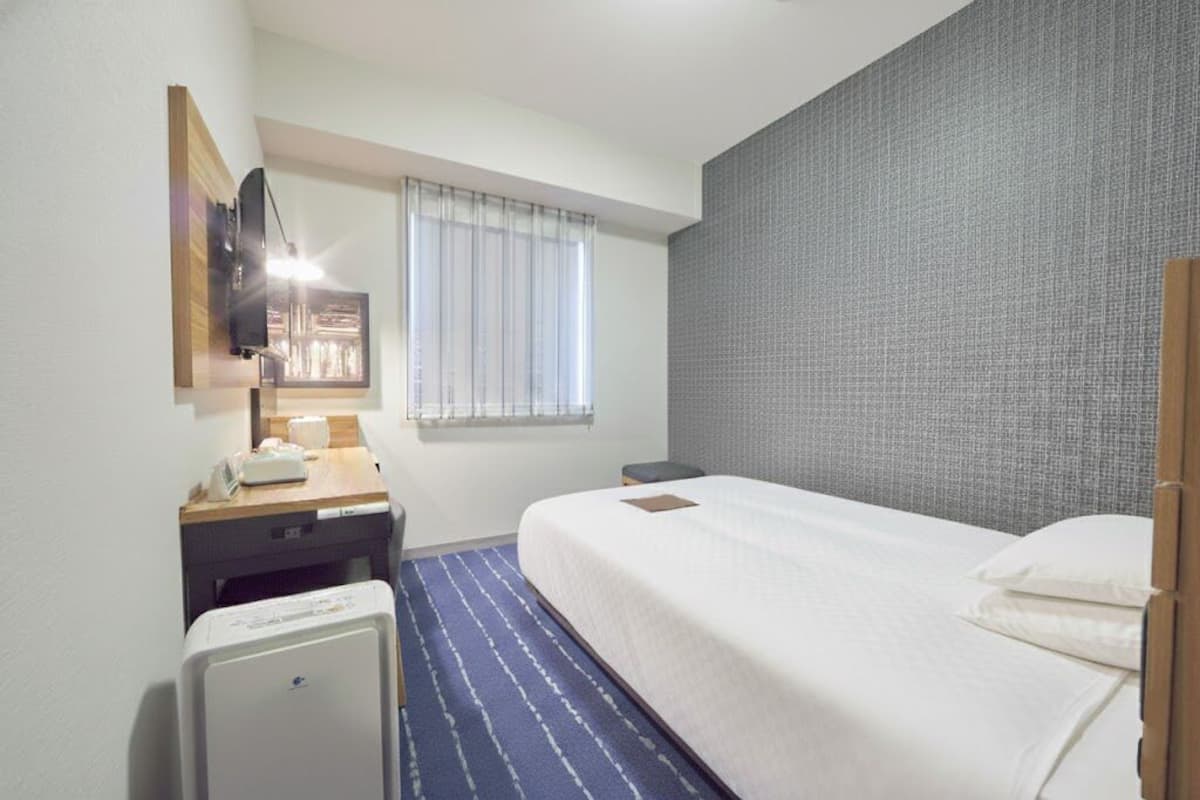 Quarto de hotel com cama, escrivaninha com luminária, aquecedor pequeno e janela com persianas.