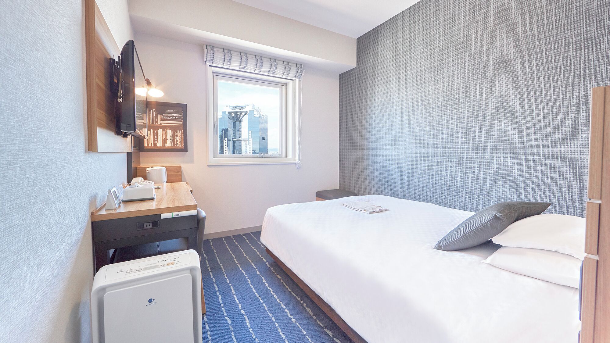 A hotel room with a bed, a desk with a telephone, a small heater, a window with a view of buildings, and a wall with a textured pattern.