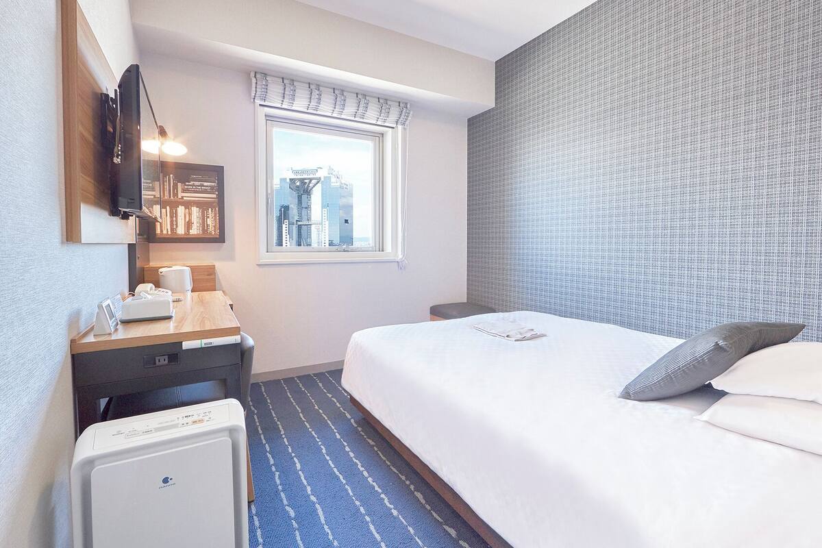 A hotel room with a bed, a desk with a telephone, a small heater, a window with a view of buildings, and a wall with a textured pattern.
