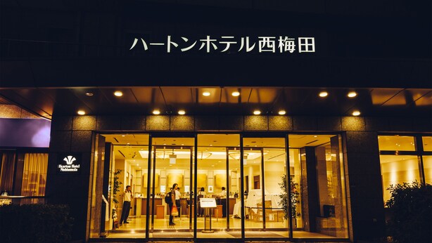 Exterior de un hotel con un letrero en japonés, entrada de vidrio y un interior bien iluminado visible a través de las puertas.