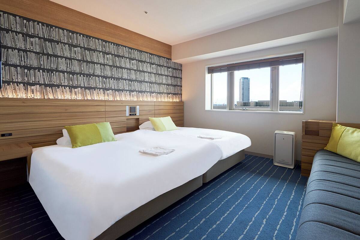 A hotel room with a large bed, a sofa, a wall-mounted heater, and a window with a city view.