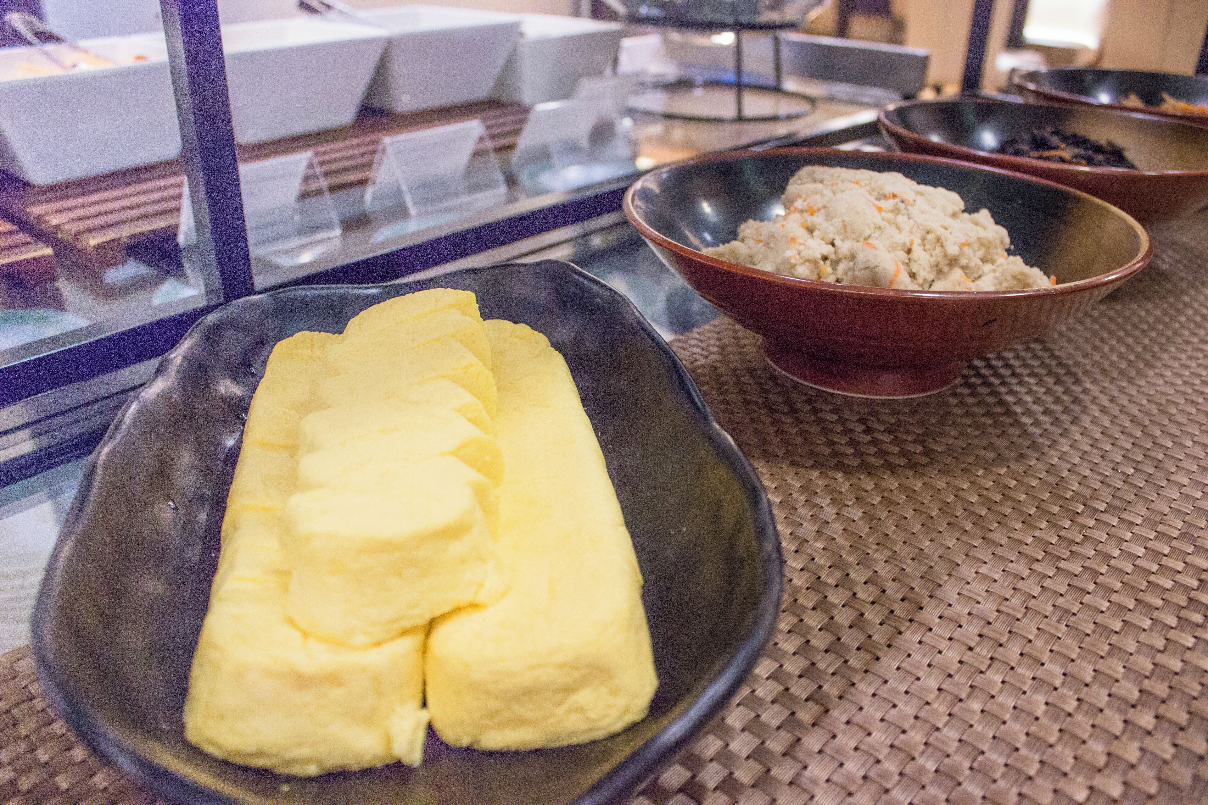 Una mesa con un posavasos tejido, un cuenco negro con comida amarilla y un cuenco marrón con comida blanca.