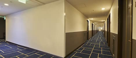 A long, narrow hotel corridor with a patterned carpet, doors on both sides, and a ceiling with recessed lighting.