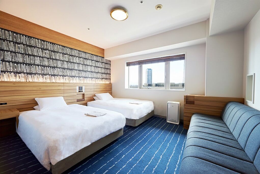 A hotel room with two beds, a sofa, a large window with a city view, and a wall-mounted artwork.