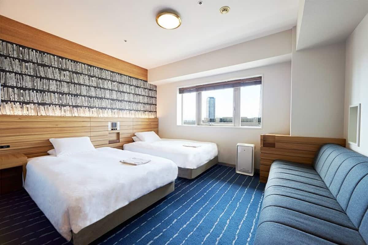 Quarto de hotel com duas camas, um sofá, uma janela ampla com vista para a cidade e uma obra de arte na parede.
