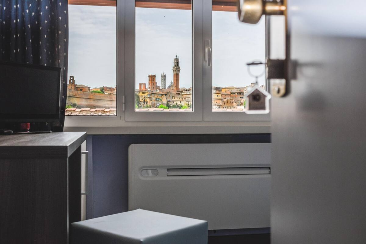 Een kamer met een raam dat uitkijkt op een stadsgezicht, inclusief een opvallende toren.