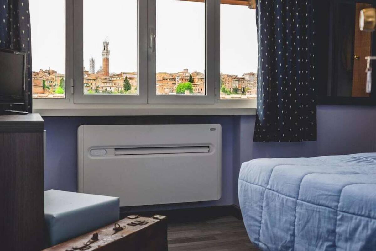 Een slaapkamer met een bed, een raam met uitzicht op een stadsbeeld, een radiator en een klein tafeltje.