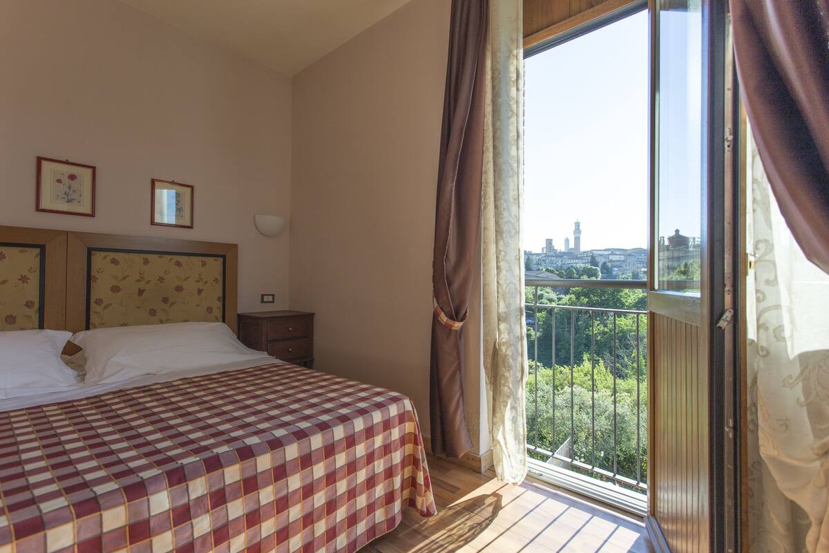 Een hotelkamer met een bed, een nachtkastje, een raam met gordijnen en uitzicht op een stadsgezicht.