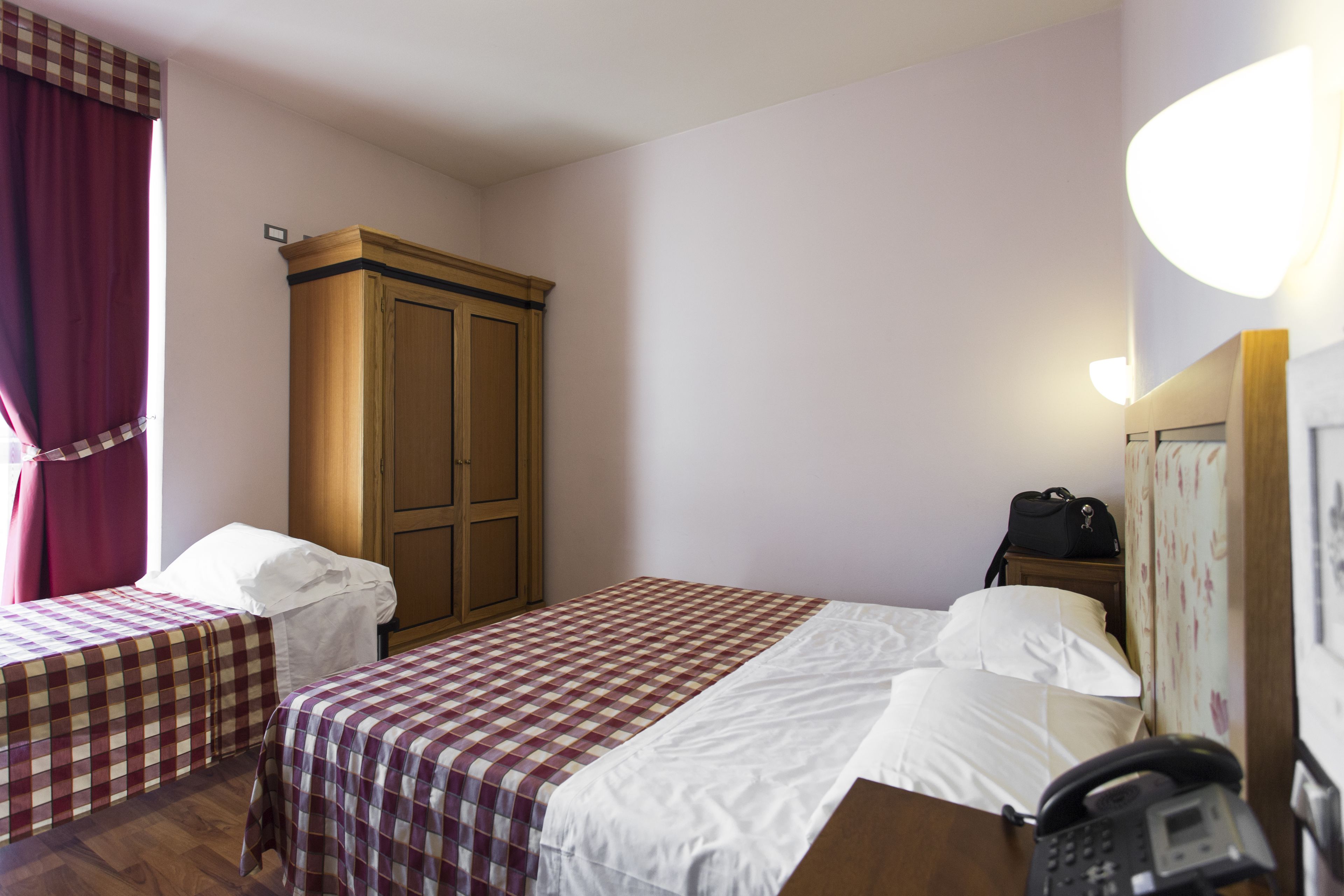 Habitación de hotel con dos camas, un armario, un escritorio con teléfono y una mesita de noche con una bolsa.