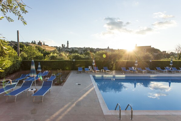 Une piscine entourée de chaises longues et de parasols, avec une végétation luxuriante et des bâtiments en arrière-plan.