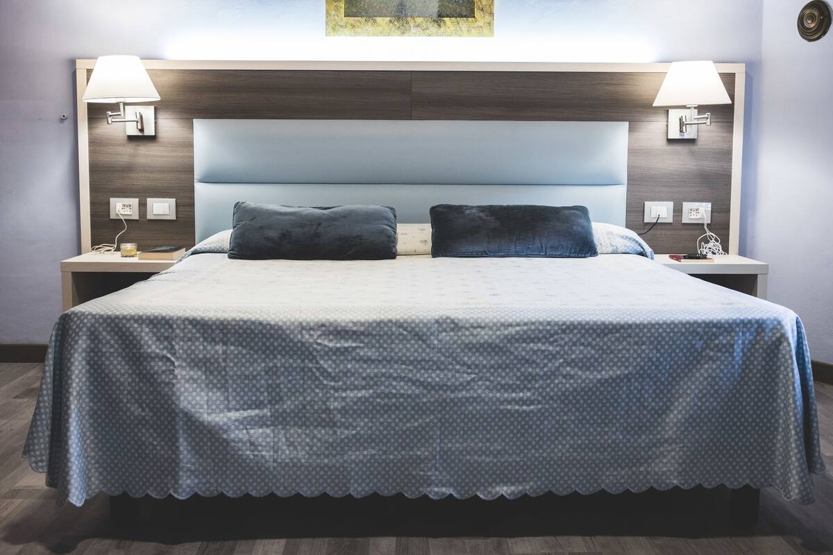 Een net opgemaakt bed met een bedsprei met patroon, twee bedlampen en een hoofdbord met een lichtblauwe panelen.