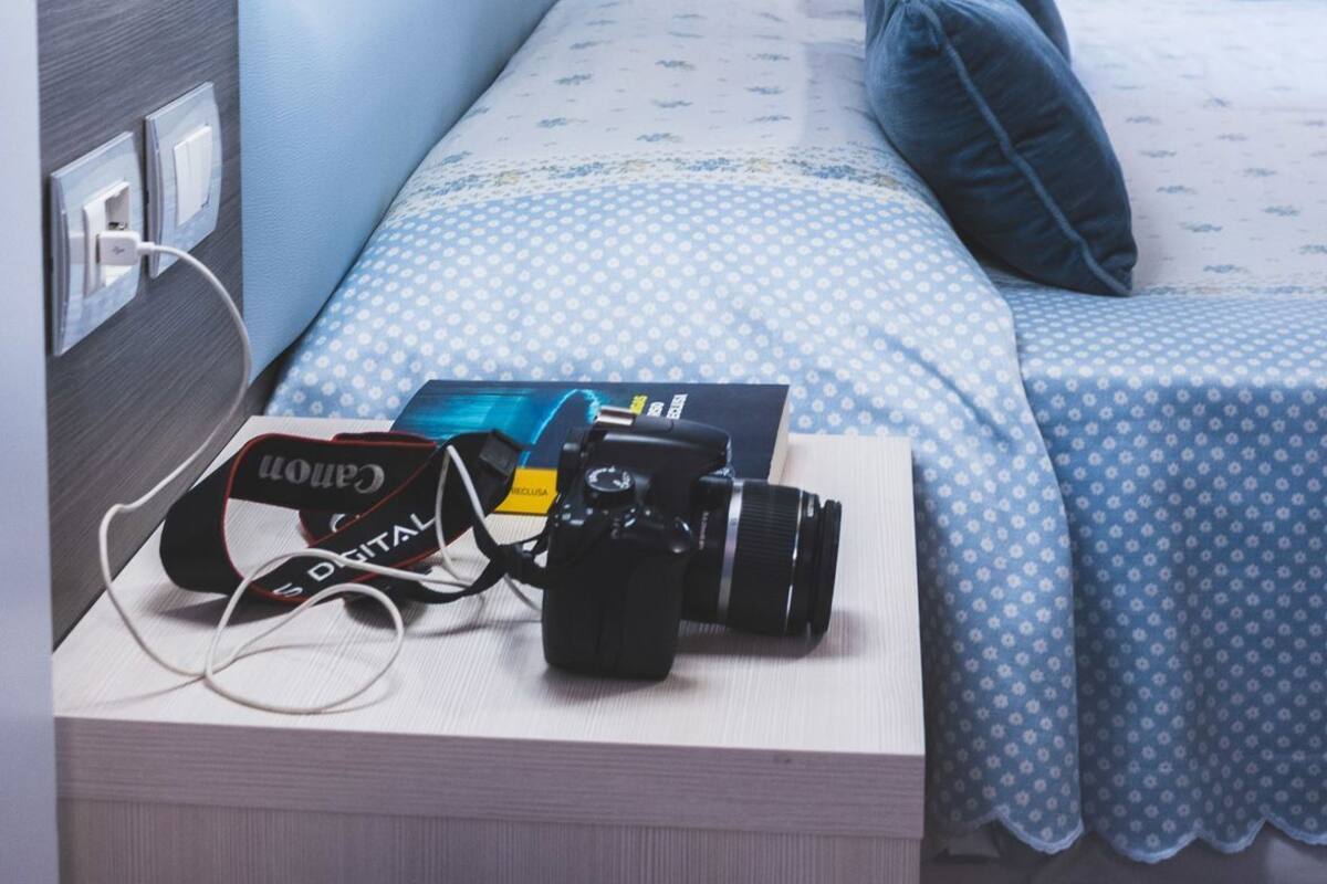Een net opgemaakt bed met een kussen, een lamp en een camera op het nachtkastje.