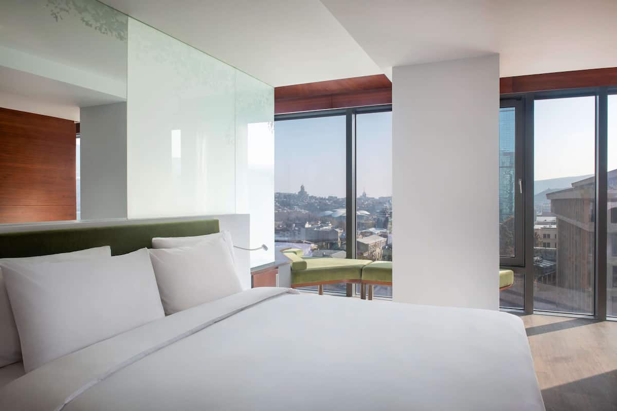 A modern hotel room with a large bed, a green chair, and a view of the city through large windows.