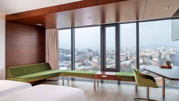 A modern hotel room with a large window offering a city view, a green sofa, a white bed, and a small desk with a chair.