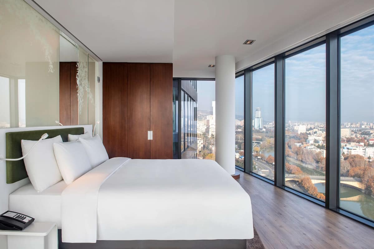 A modern hotel room with a large bed, a city view, and a glass wall.