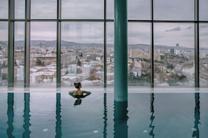 A large indoor swimming pool with a view of a cityscape through large windows.