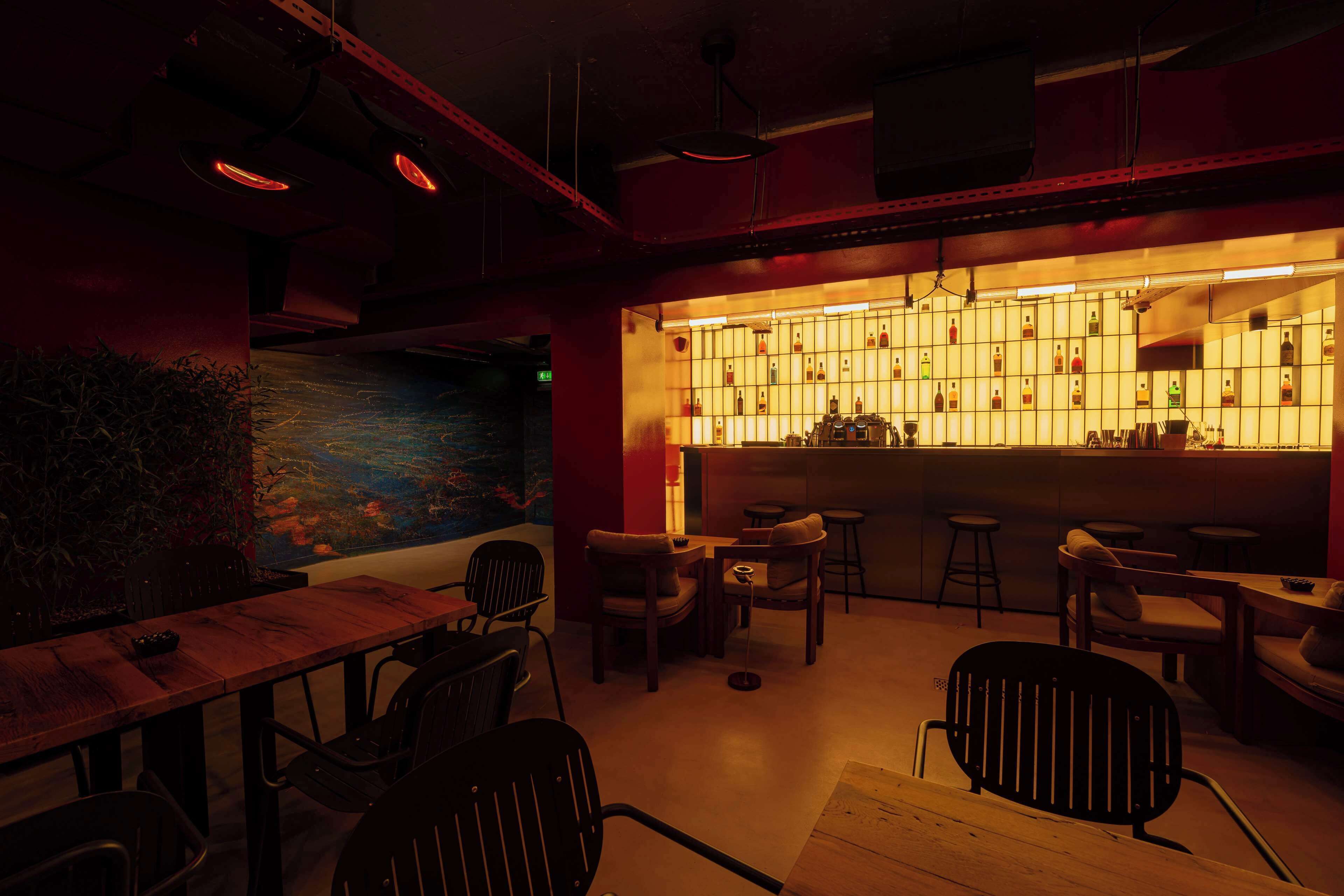 Zona de bar tenuemente iluminada, con mobiliario de madera y una pared de botellas iluminadas.
